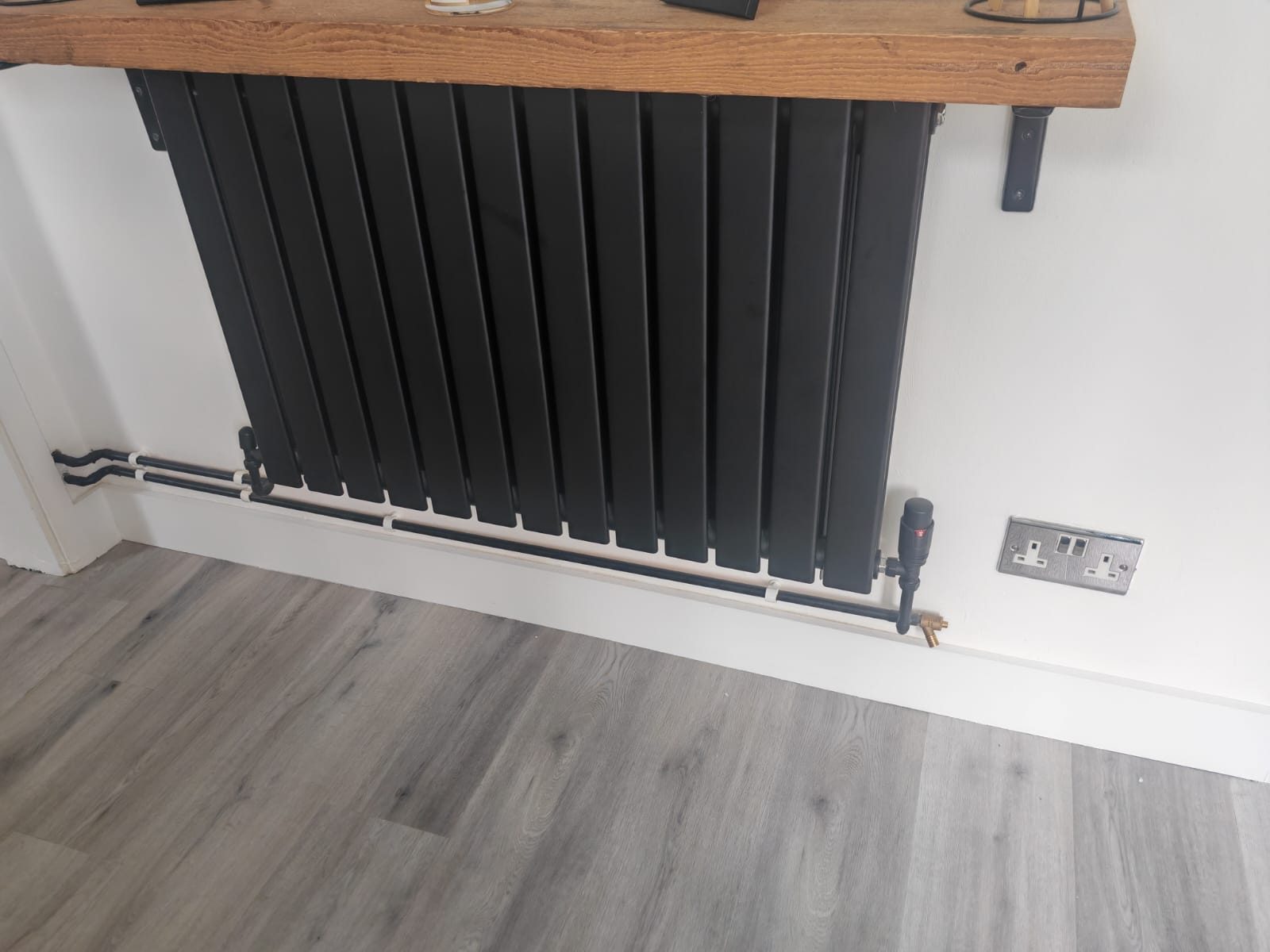 Black flat-panel designer radiator under a solid wood shelf with neat pipework